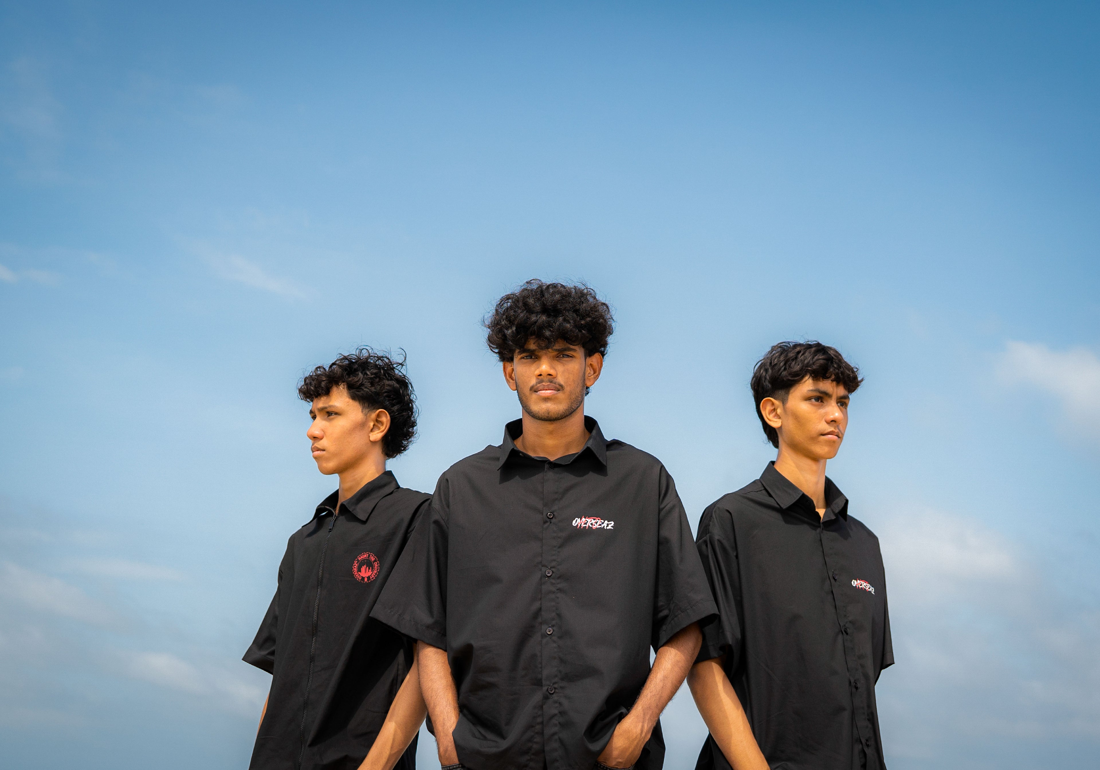 Three individuals wearing black shirts with logos against a blue sky.